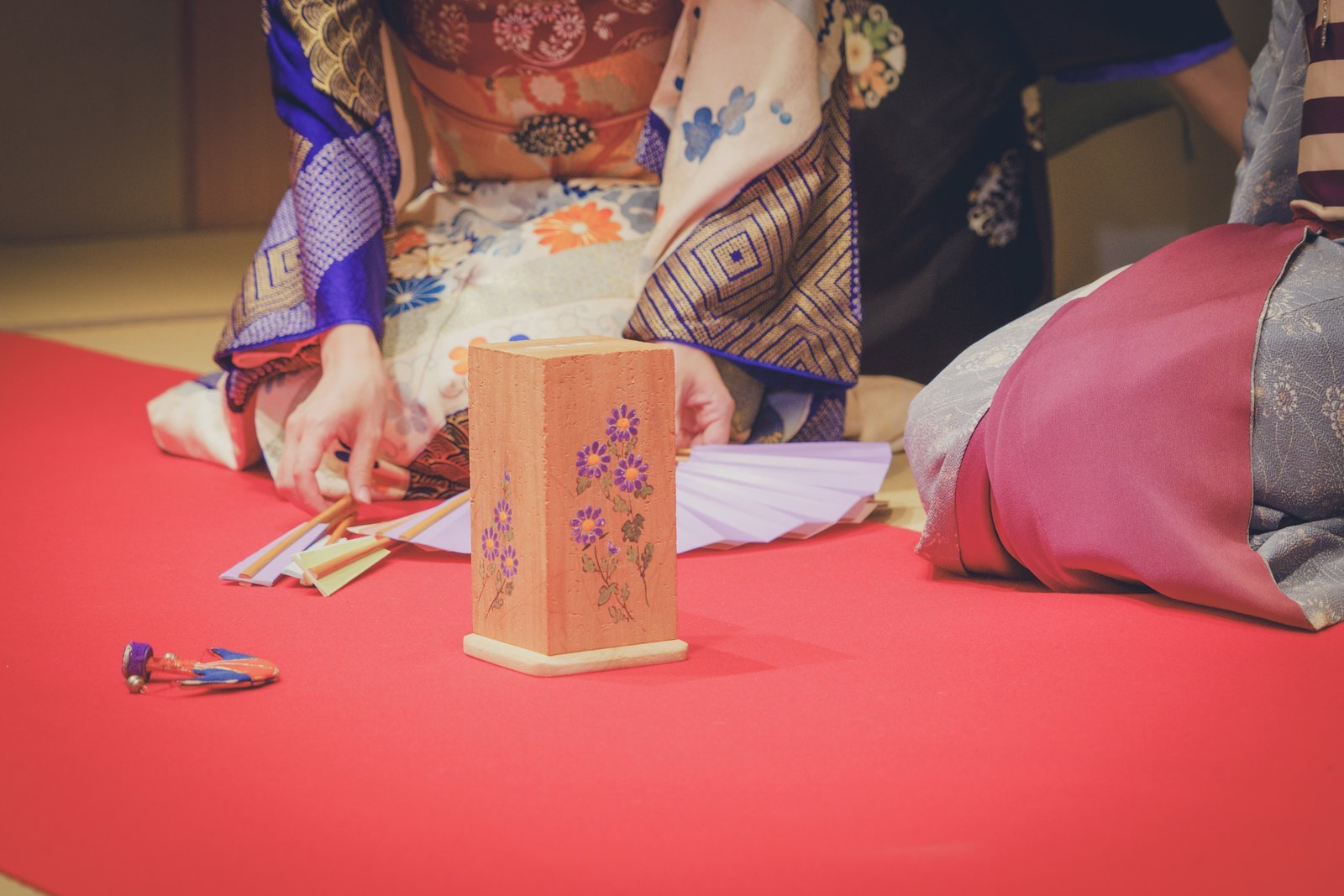 The makura stand, butterfly-shaped cho, and tosenkyo fans laid out on red felt at a Miyakodori ozashiki, with geisha seated around them