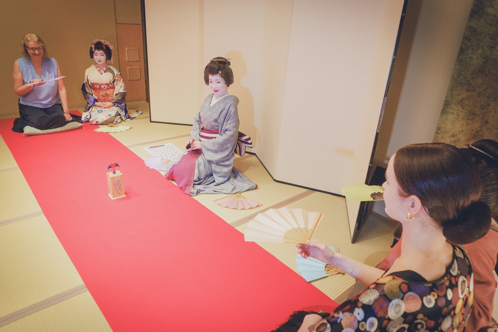 A tosenkyo match at Miyakodori — guests holding fans on either side of the red felt, a geisha seated by the makura to read and call each score