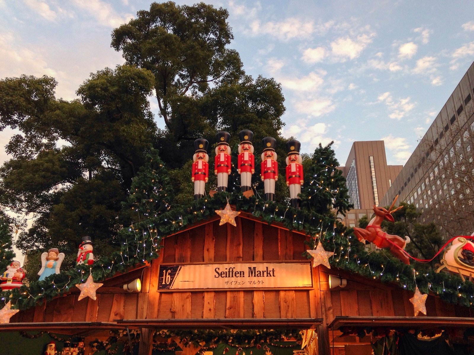 A wooden Christmas market hut decorated with a nutcracker, a wreath with star lights, and a reindeer and sleigh, against a dusk sky