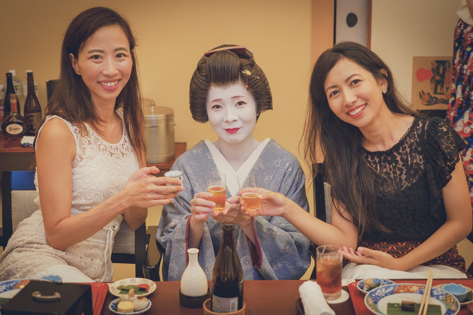 A geisha and two international guests raising a toast at a low table in a private tatami room, under warm lighting