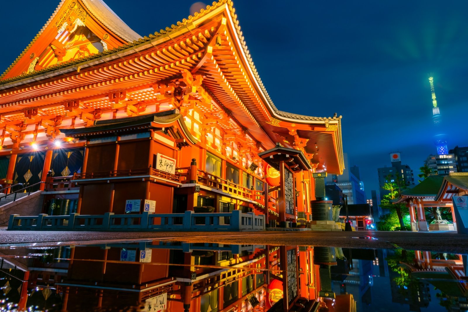 Senso-ji's Main Hall illuminated at night, reflected in water, with Tokyo Skytree rising in the background
