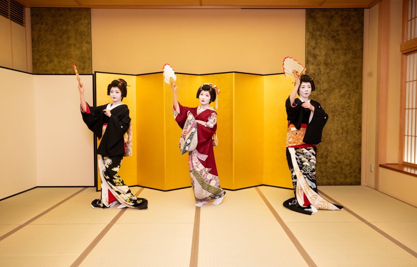 Three Asakusa geisha dancing nihon buyo with fans in front of a gold folding screen at a private ozashiki