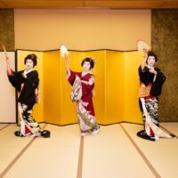 Three Asakusa geisha dancing nihon buyo with fans in front of a gold folding screen at a private ozashiki
