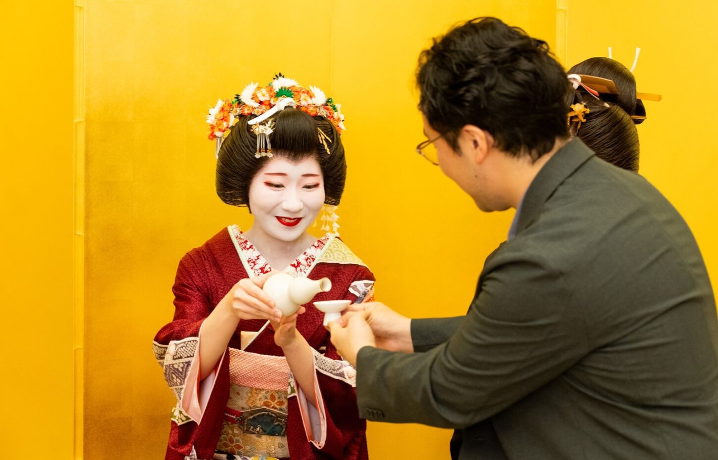 An Asakusa geisha pouring sake for a guest in front of a gold folding screen, the setting where her fee is earned