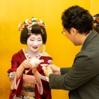 An Asakusa geisha pouring sake for a guest in front of a gold folding screen, the setting where her fee is earned
