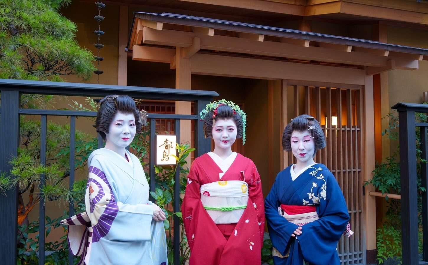 An Asakusa hangyoku in a red furisode between two senior geisha at the entrance of a teahouse