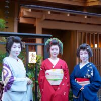An Asakusa hangyoku in a red furisode between two senior geisha at the entrance of a teahouse