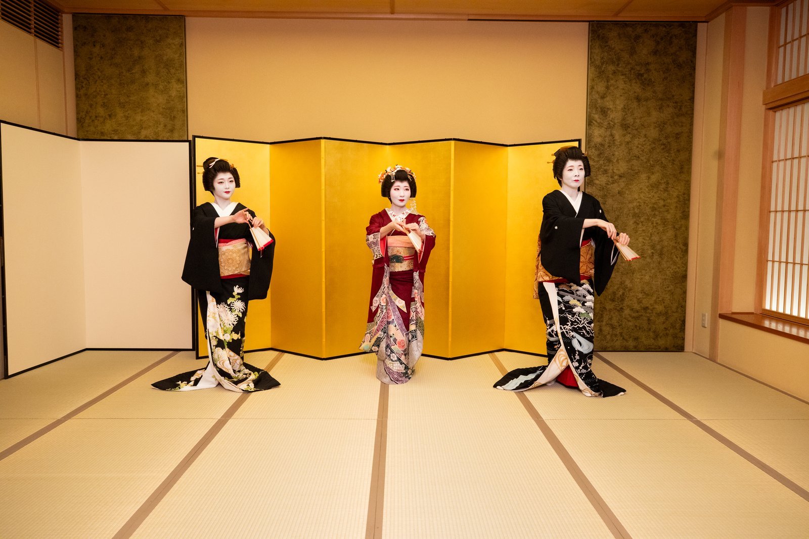 Three Asakusa geisha in formal kimono before a gold screen, two in black kimono with trailing hems