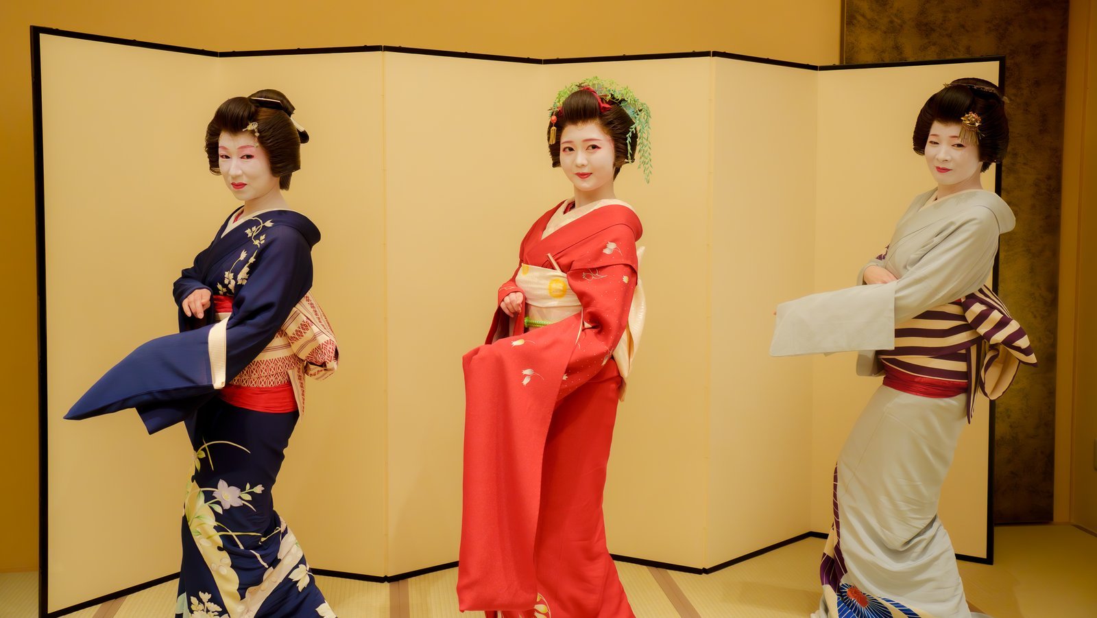 An Asakusa hangyoku in a red furisode between two full geisha before a gold screen