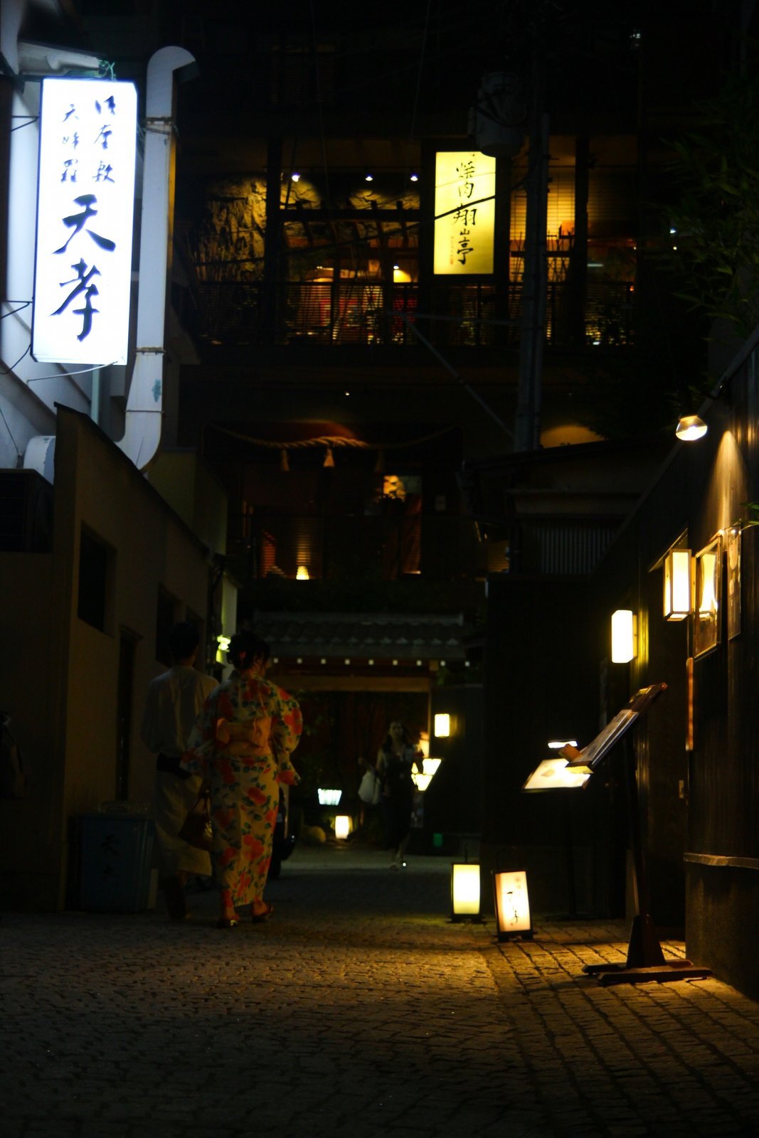 A woman in kimono walking down a lantern-lit alley in a geisha district at night