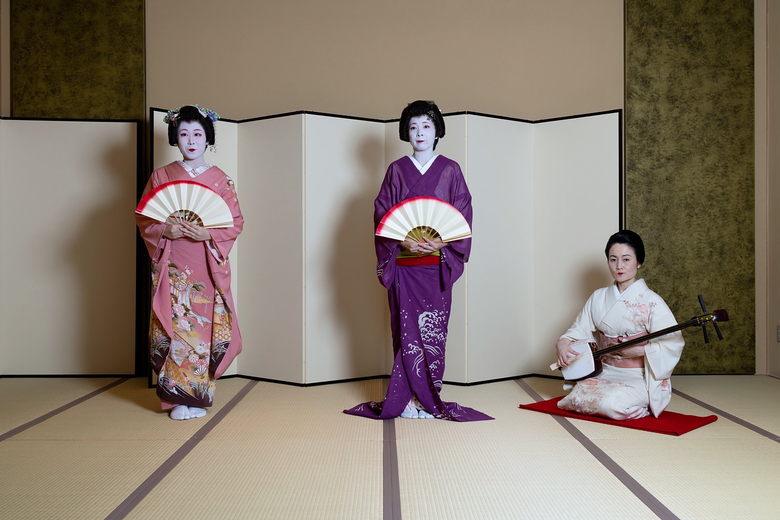 Two Asakusa geisha with fans and a jikata with a shamisen in front of a gold screen