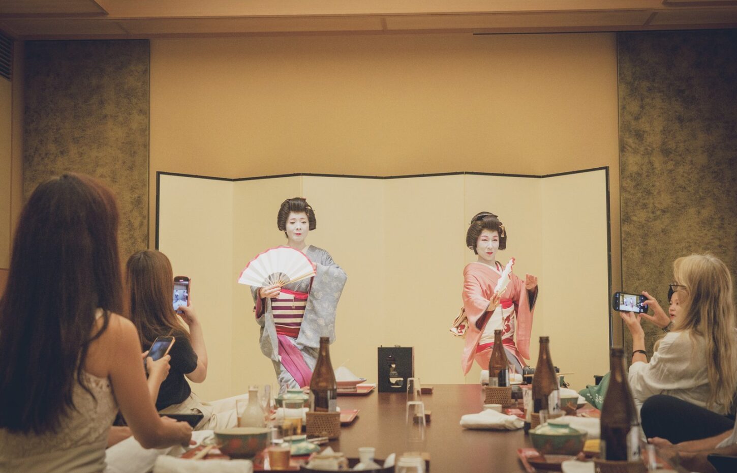 Two geisha dancing side by side in front of a gold folding screen while guests at the banquet table take photos