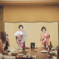 Two geisha dancing side by side in front of a gold folding screen while guests at the banquet table take photos