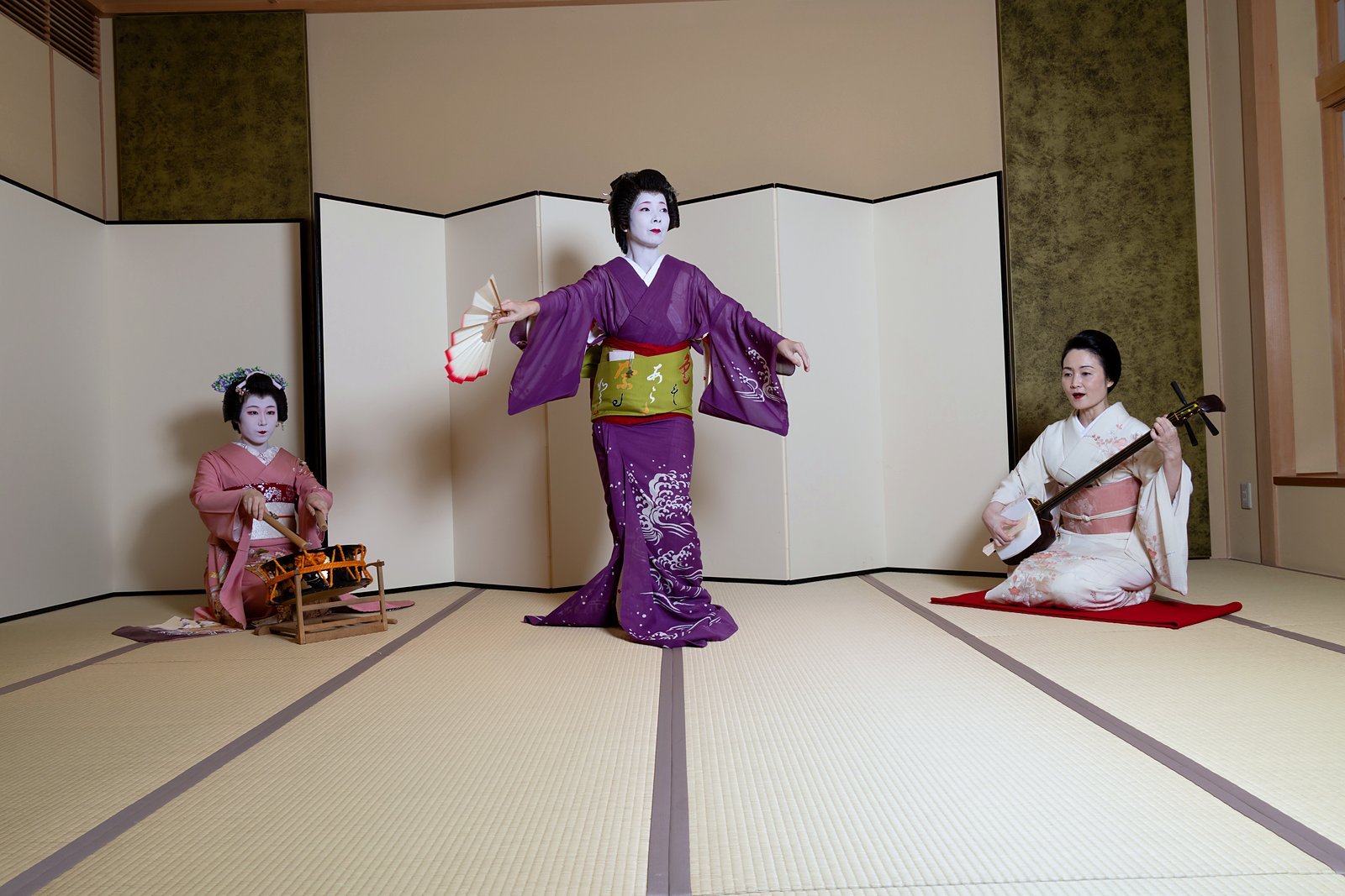 Three geisha on the tatami: one dances with a fan in the centre, one plays a drum and one plays the shamisen