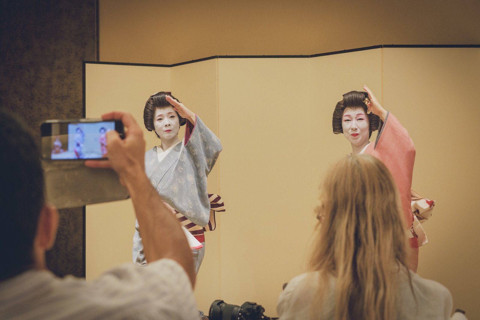 Two geisha in grey and pink kimono dancing the same movement together, as guests film them