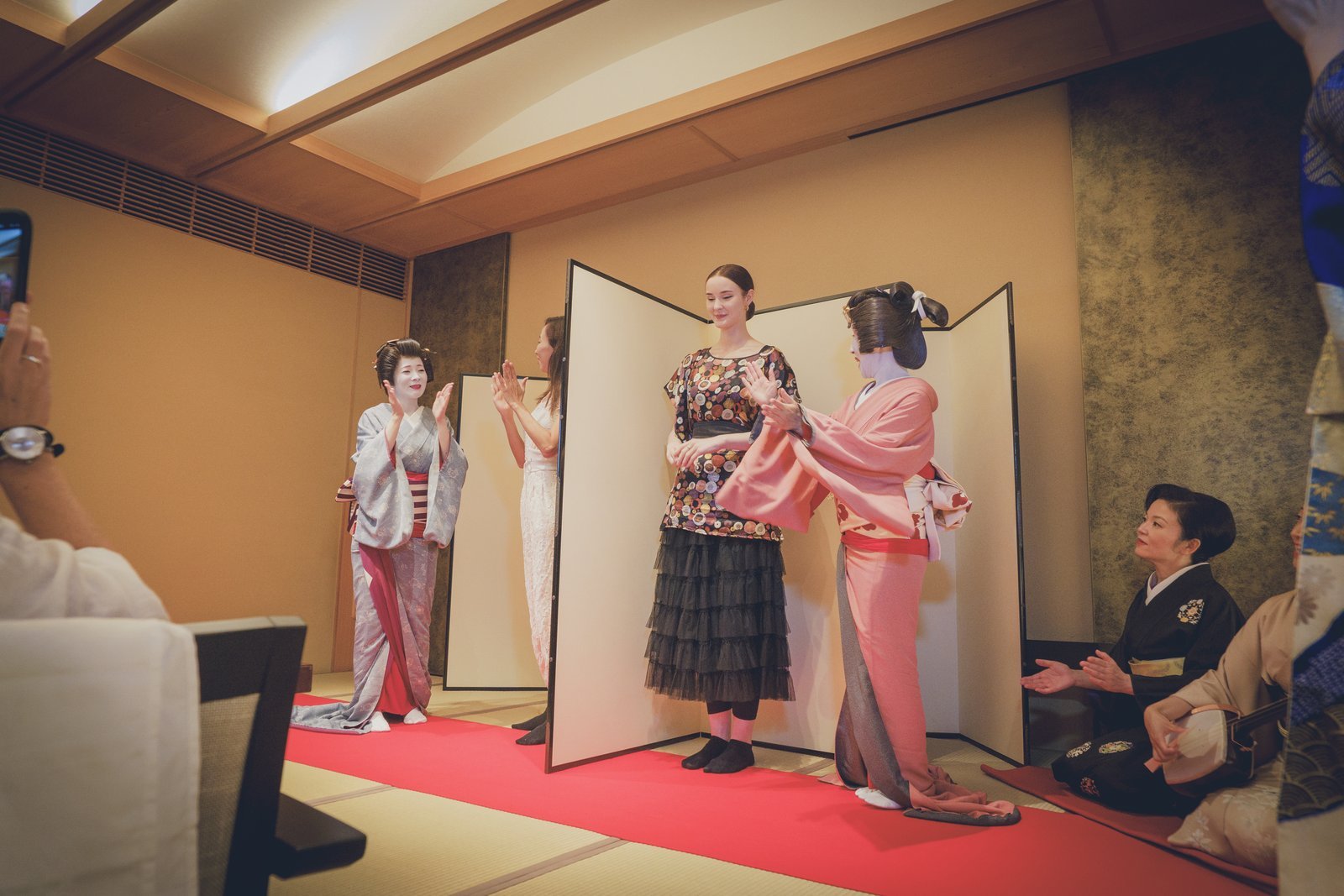 A guest playing tora tora beside a folding screen, with a geisha on each side and a shamisen player to the right
