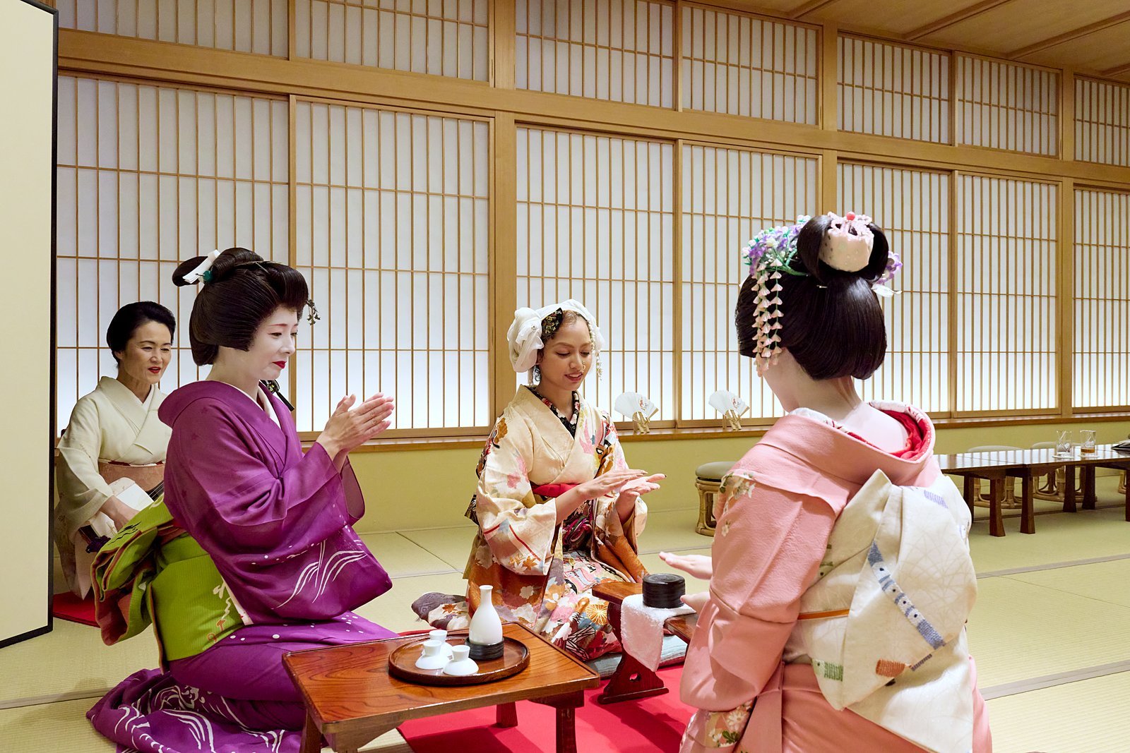 A guest playing konpira fune fune with a geisha across a small stand, while another geisha claps along and a shamisen player sits behind