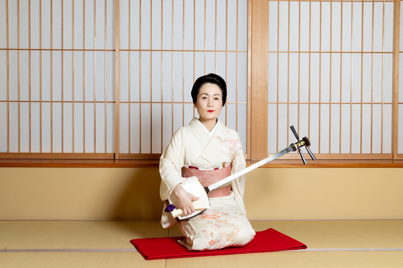 A jikata in a cream kimono kneeling on a red cloth with her shamisen, in front of shoji screens