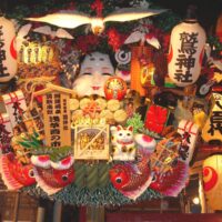 A large kumade rake with an Okame mask and sea bream, under Otori Shrine lanterns at night